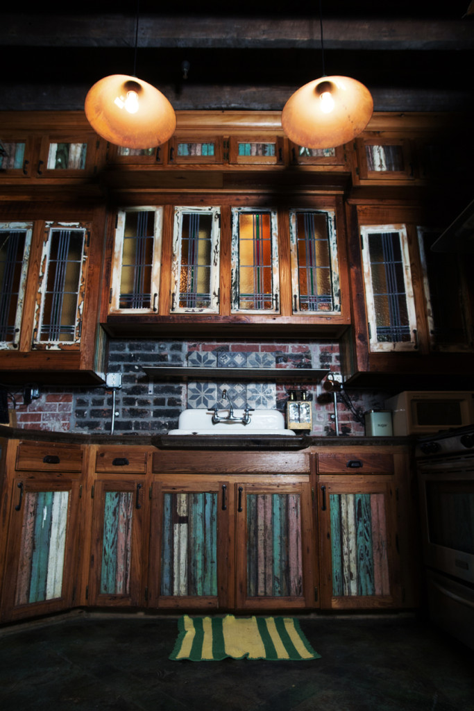 In the kitchen, a porcelain sink is offset by a brick and concrete backsplash. The upper cabinet doors are stained glass, while the lower cabinet doors are covered with multicolor wood panels. A butcher block cutting board is built into the countertop.