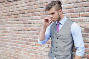 casual young man standing next to a brick wall with a cigarette in his mouth