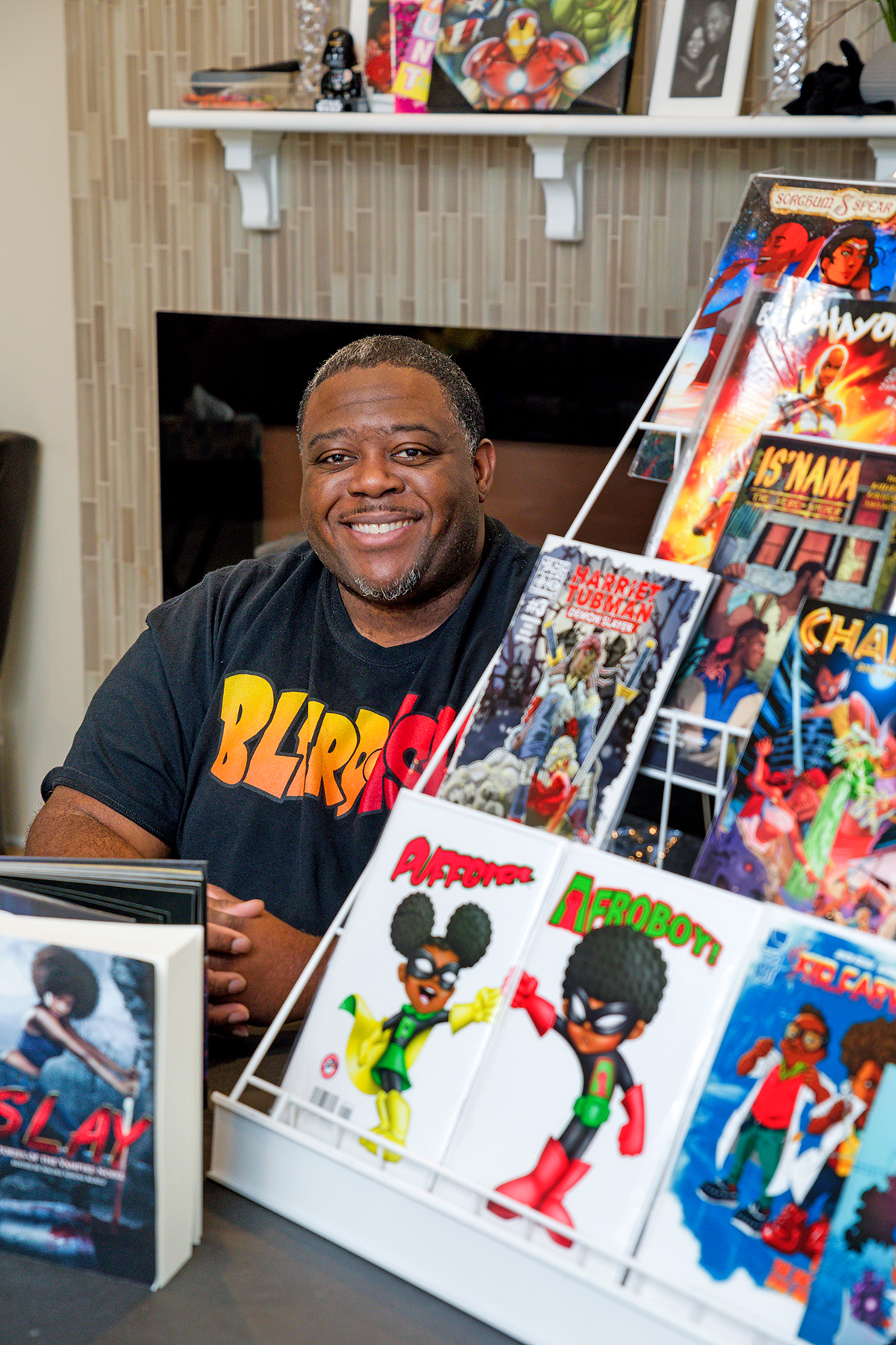 Keith Cooper of Blerdish Sits in front of a fireplace with a display of comic books to his left.