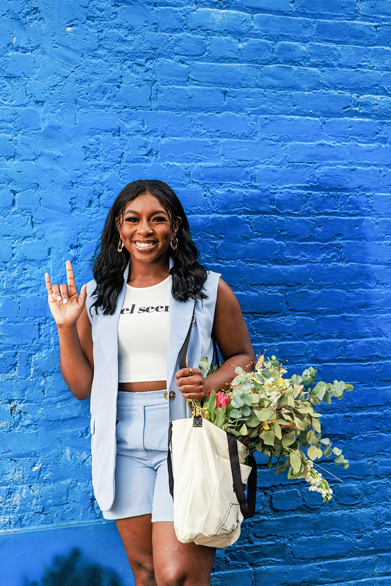 Feel Seen founder Tracee Albert signing “I love you” in American Sign Language. Standing in front of a bright blue brick wall.