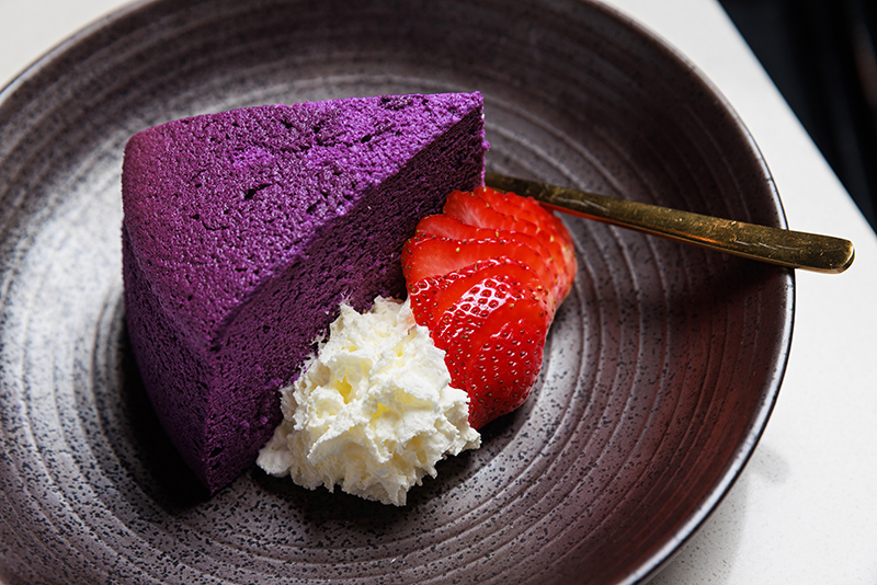 Slice of the bright purple, Ube Cheesecake, with whipped cream and sliced strawberry on the side